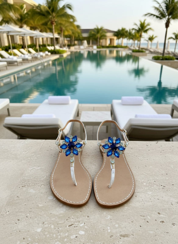 Pair of Ankalia sandals in white leather with blue flower embellishments poolside.