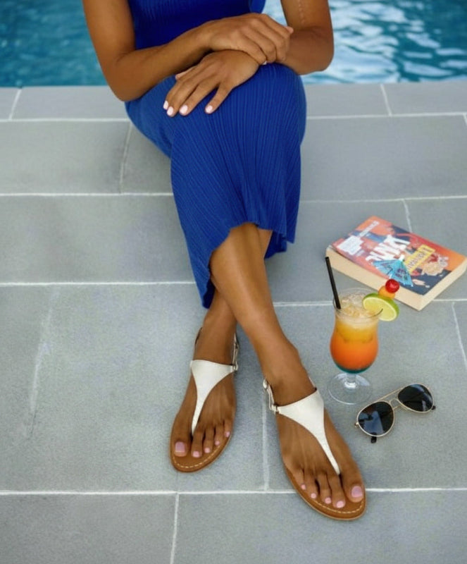 Person in a blue dress sitting by a pool with a drink and sunglasses on the ground.