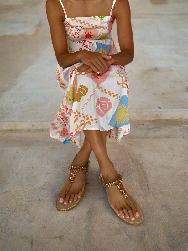 Person wearing a colourful summer dress and tan flat sparkly sandals sitting on a footpath.