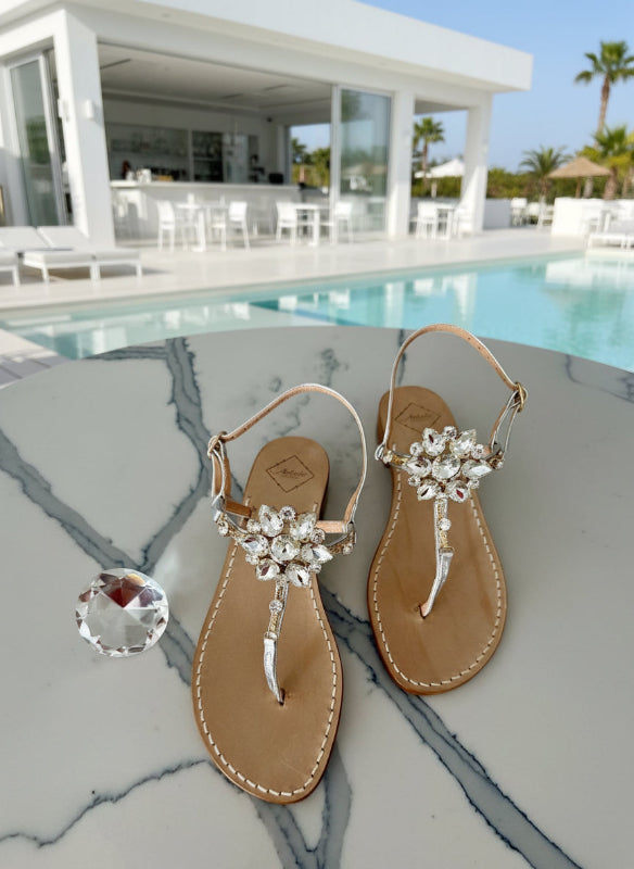 Pair of silver sandals with Swarovski crystal embellishments on a marble table poolside.